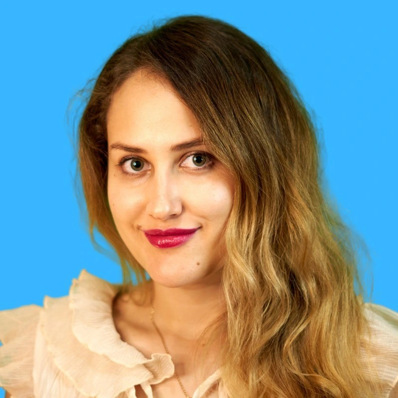 Woman with long curly blonde hair, wearing a cream top, smiles against a bright blue background.