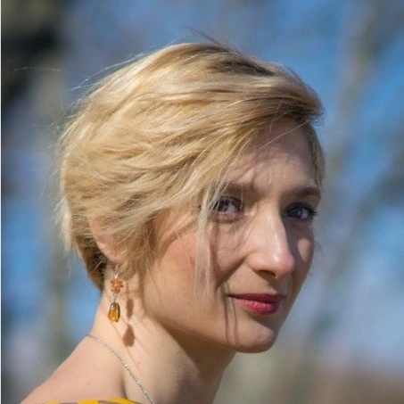 Portrait of a person with short blonde hair, wearing a yellow top and earrings, standing outdoors in bright daylight with a blurred natural background.