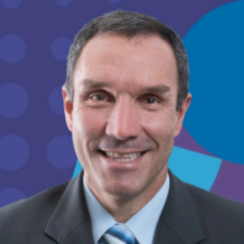 Smiling man in a suit against a blue and purple patterned background.